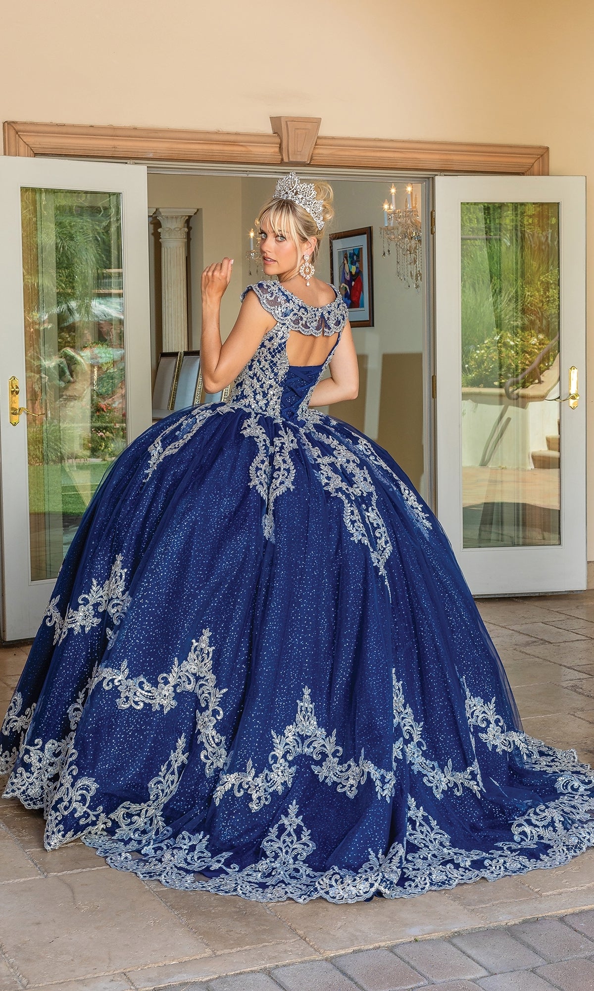 A woman outdoors wears the Navy Blue Quinceanera Dress 1709 with lace embroidery and a tiara, turning toward the camera. Glass doors and a chandelier appear in the background.